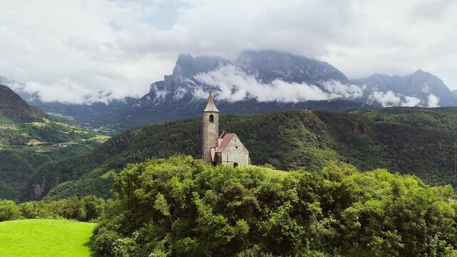 Chapel on green hill in Dolomites, with forest and mountain background, aerial view. South Tyrol, Italy