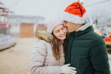 Naklejka premium Young couple in winter coats share a tender kiss while one partner wears a Santa hat. The affectionate moment is captured at a festive outdoor fair during the holiday season.
