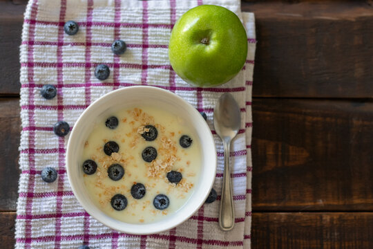 A healthy breakfast or snack consisting of a bowl of yogurt with blueberries and a green apple on a wooden table