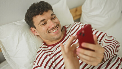 Man in striped pajamas lying on bed, smiling, waving and tapping a red smartphone in bedroom;...