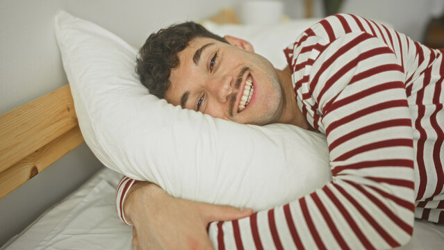 Man hugging a white pillow on bed in building, puckers lips with closed eyes and smiles while wearing red and white striped pyjama and leaning against wooden headboard; morning contentment.