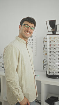 Young man wearing glasses, smiling and turning his head toward an eyeglass display and eyechart in a building; confidence style choice.