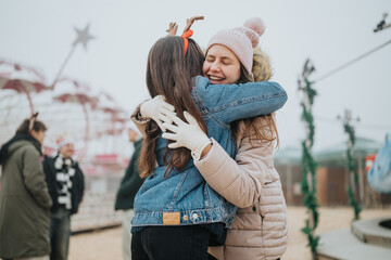 Naklejka premium Two young women embrace outdoors at a winter fair wearing warm coats and gloves. The friends hug and smile amid a festive amusement-park atmosphere.