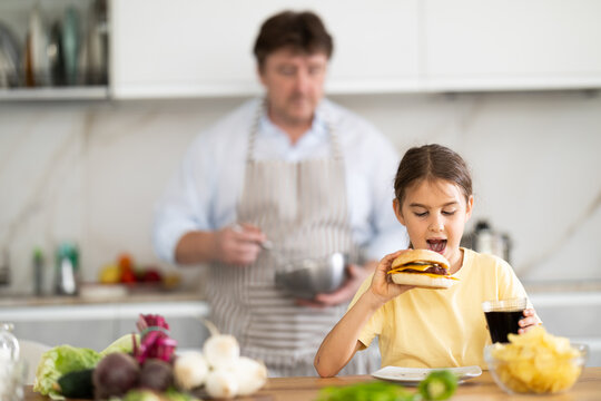 Little daughter eats meat burger and drinks carbonated drink in the kitchen. In background, father prepares healthy vegetable salad