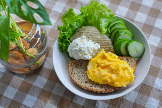 Healthy breakfast with sourdough bread topped with creamy scrambled eggs, served with a side of rich Greek yogurt, fresh sliced cucumbers, green lettuce, and grilled chicken breast on a table.