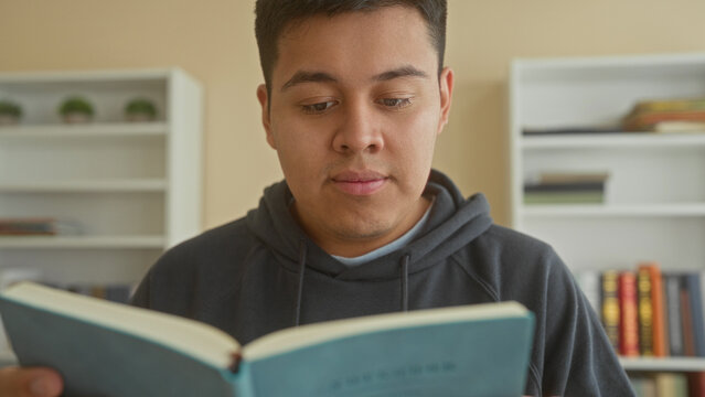 Man holding open book and reading with focused gaze in a shelf lined library, face visible and hands supporting the pages; quiet concentration.