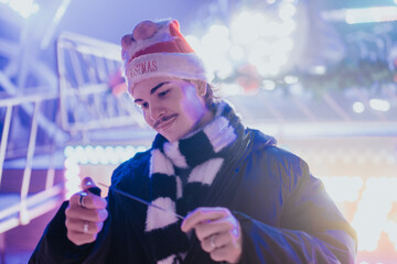 Naklejka premium A young man wearing a Santa hat and scarf adjusts a string of lights at a festive nighttime market.