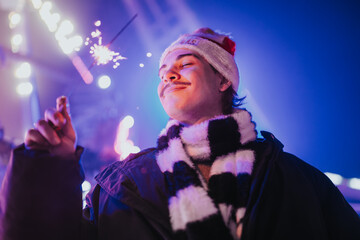 Naklejka premium Young man wearing a Santa hat and striped scarf holds a lit sparkler at night. Festive nighttime portrait captures a smiling friend enjoying holiday lights.