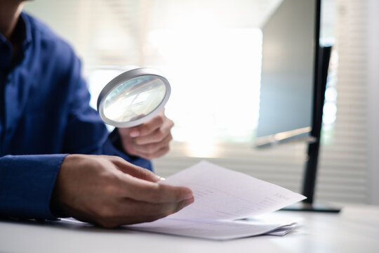 Business professional inspects financial documents with a magnifying glass at an office desk. The scene represents audit review, accounting verification, compliance, risk assessment and analysis.