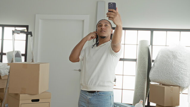 Man in white polo holds smartphone aloft taking a selfie among moving boxes inside an apartment building; new home optimism.