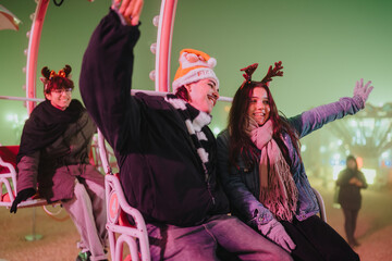 Naklejka premium Three friends wearing winter hats and antlers smile and wave on a carnival ride at night. A festive holiday scene with colorful lights and joyful expressions at an outdoor amusement area.