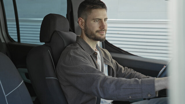 Man driving with hands on steering wheel inside car beside corrugated metal building facade, gazing forward in a steady pose; calm contemplation.