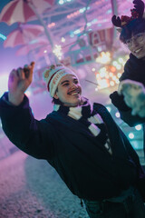 Naklejka premium Two friends wearing winter hats and scarves smile while holding sparklers at night. A festive carnival scene glows with colorful lights and joyful atmosphere.