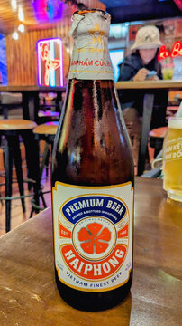 A cold bottle of Haiphong Premium Beer sits on a wooden table in a dimly lit bar with neon lights in the background