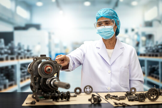 Factory engineer in protective mask and hairnet inspecting a mechanical engine part with gears and tools on a workbench.