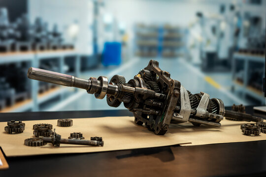 Disassembled manual transmission gearbox with shaft, gears and metal parts on a workshop table, with a blurred automotive repair shop in the background.