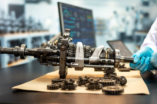 Technician wearing blue gloves inspects a disassembled vehicle gearbox with metal gears on a workbench in a modern repair workshop.