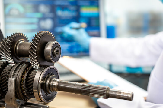 Close view of metal gearbox gears and transmission shaft on a workbench, with a mechanic using a computer in a bright automotive repair workshop