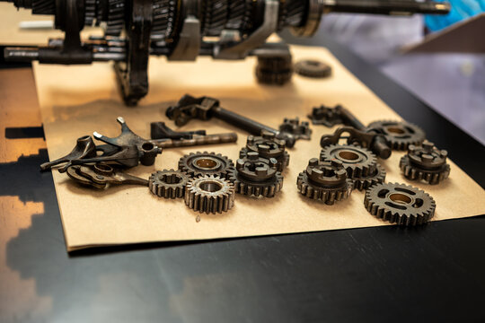 Close-up of assorted metal gears, chains and small machine parts laid on cardboard beneath industrial equipment in a workshop.