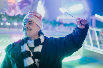 Naklejka premium Young man wearing a Santa hat and scarf smiles while holding a lit sparkler. A festive portrait of friends enjoying holiday lights at a nighttime fair.