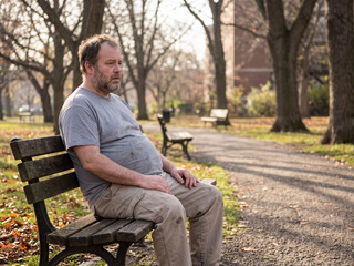 Obraz premium Middle-aged heavyset man sitting on wooden park bench in urban park