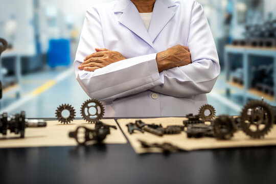 Confident engineer or technician in a white lab coat standing with folded arms behind a workbench with metal gears and machine parts in a modern workshop.