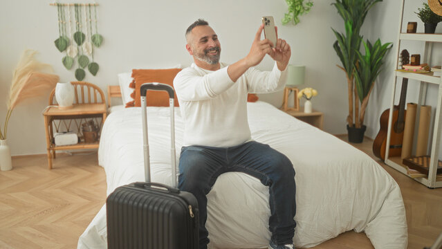 Man seated on bed with carryon suitcase, holding smartphone with both hands taking a selfie in a bedroom inside a building; anticipation travel.