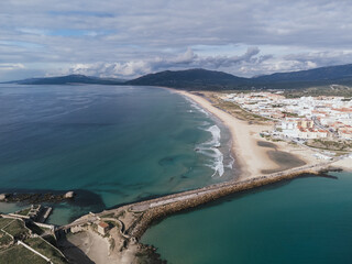 Aerial view of a long sandy beach curving along turquoise ocean waters beside a coastal town with white buildings and mountains in the distance in Tarifa, Spain © Felix Morion