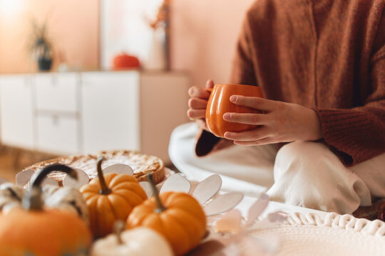 Cozy woman holding a warm orange mug beside small pumpkins and autumn decor in a softly lit home.