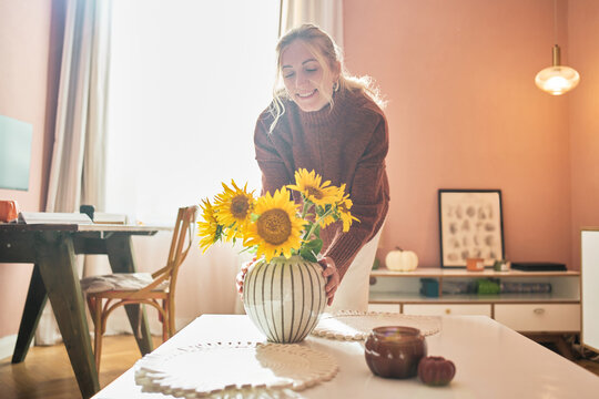 Smiling woman arranging bright sunflowers in a striped vase on a table in a warm, sunlit living room.