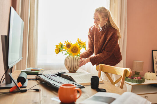 Smiling woman arranging sunflowers in a vase on a home office desk with computer, books, coffee and papers in warm morning light.