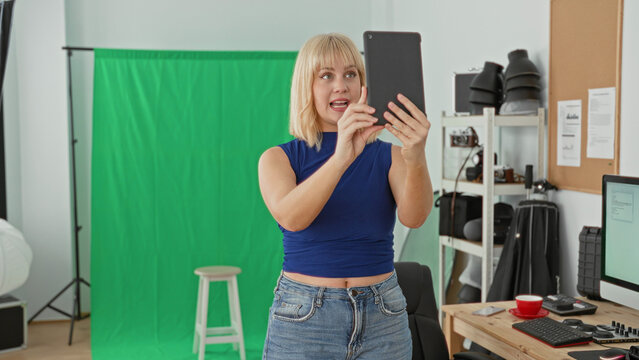 Woman holding tablet with both hands, smiling and looking at screen in a studio with green screen and stool; content creation confident.