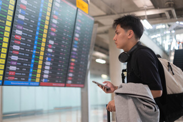 Young traveler checking a flight information board at an airport while holding a smartphone, wearing headphones and carrying a backpack, representing travel planning, departure, adventure, and journey © Bordinthorn
