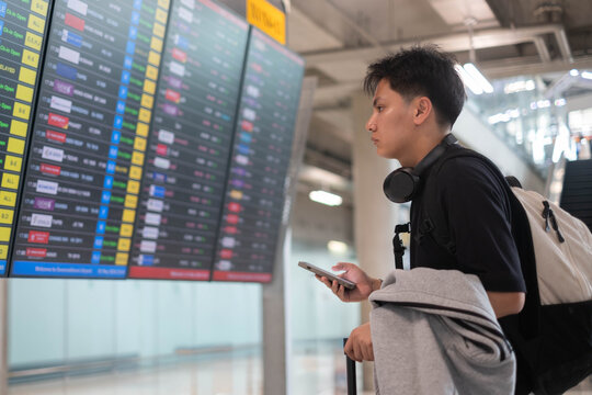 Young traveler checking a flight information board at an airport while holding a smartphone, wearing headphones and carrying a backpack, representing travel planning, departure, adventure, and journey