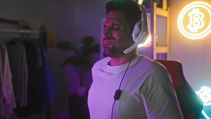 Man gamer wearing headset rubs shoulder and grimaces in a streaming studio, seated at computer with neon signs  strain discomfort. © Krakenimages.com