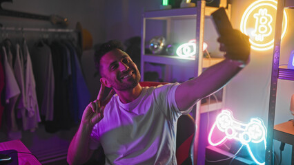 Man in white t shirt taking a selfie with smartphone, making shaka sign with his hand amid neon gamepad and bitcoin lights in a streaming studio  playful social gaming. © Krakenimages.com