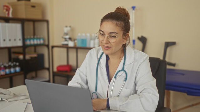 Young woman doctor claps hands at laptop in clinic while wearing stethoscope and glasses; joy accomplishment.