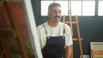 Man painter in apron speaks while gesturing with open hand toward canvas and easel in bright studio  focused creativity. © Krakenimages.com