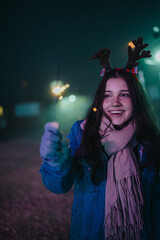 Naklejka premium A smiling teenage girl wearing reindeer antlers holds a lit sparkler at night. She celebrates outdoors with friends in a festive winter scene.