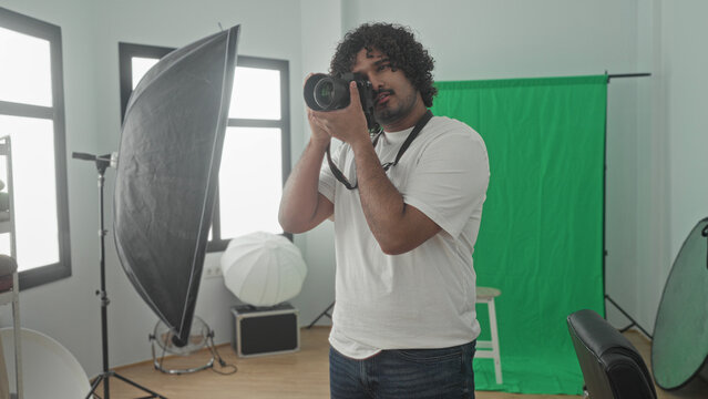 Man photographer holding camera with strap around neck, adjusting lens and looking through viewfinder beside green screen, softbox and stool in studio; focused creativity.