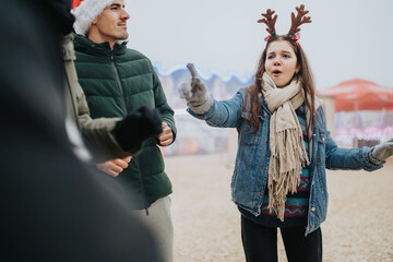Naklejka premium Teen girl wearing reindeer antlers and a scarf gestures while singing with friends outdoors. A festive winter market scene with friends enjoying holiday fun and music.