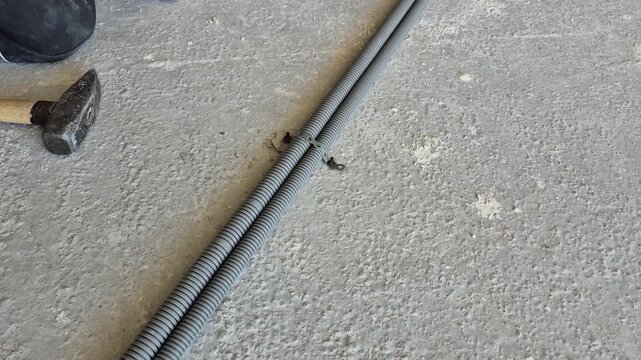 Top-down view footage of an electrician using a hammer and a nail-in anchor to secure a clamp to the floor, fastening the electrical conduit pipe.