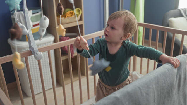 Little blond boy reaches right hand to hanging plush mobile over wooden crib inside building nursery; gentle curiosity.