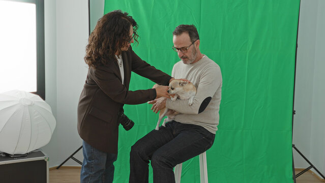 Man holding small dog while woman photographer adjusts the pet and touches his shoulder in studio; focused teamwork.
