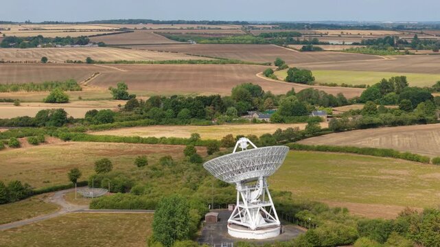 Aerial view of a massive white radio telescope dish for interstellar astronomy research surrounded by green fields in Cambridgeshire, UK