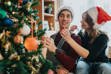Naklejka premium A young couple wearing Santa hats decorates a Christmas tree in a cozy home. The smiling pair hangs ornaments and tinsel together in a festive holiday scene.