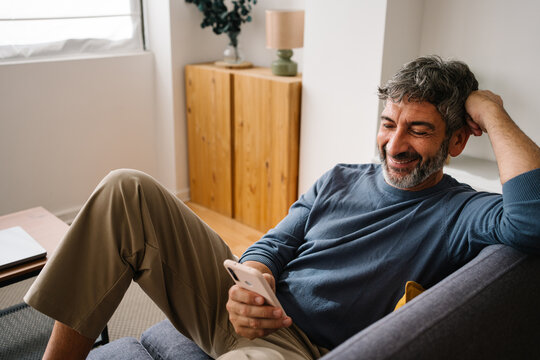 Relaxed middle aged man lounging on sofa using smartphone app in cozy living room at home