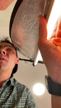 low-angle studio view of man positioning circular silver reflector beside illuminated fluorescent tube, looking upward as light reflects across shiny metallic