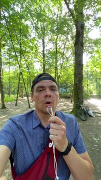 man in cap holds white earbud cable and face mask near mouth for contemplative selfie among tall trees with bright green canopy in sunlit forest during daylight