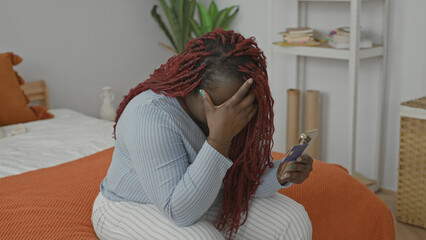 Young african american woman sitting on bed in bedroom covering her face with her hand while holding smartphone  sadness isolation. © Krakenimages.com